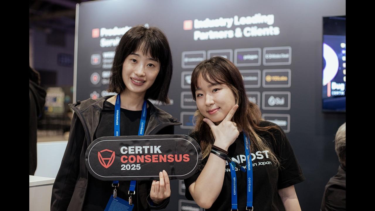 Two attendees holding a CertiK Consensus 2025 sign at the CertiK booth