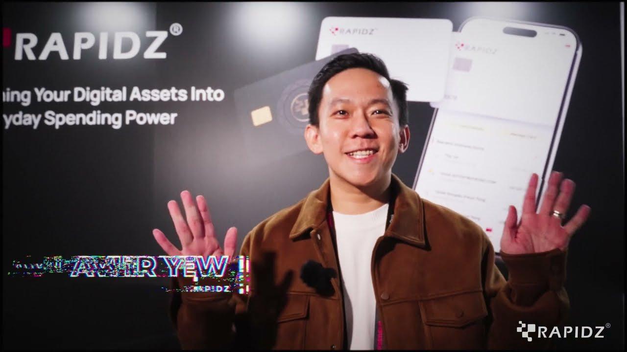 A RapidZ team member waving both hands in front of the RapidZ card and app display at their Consensus Hong Kong 2026 booth